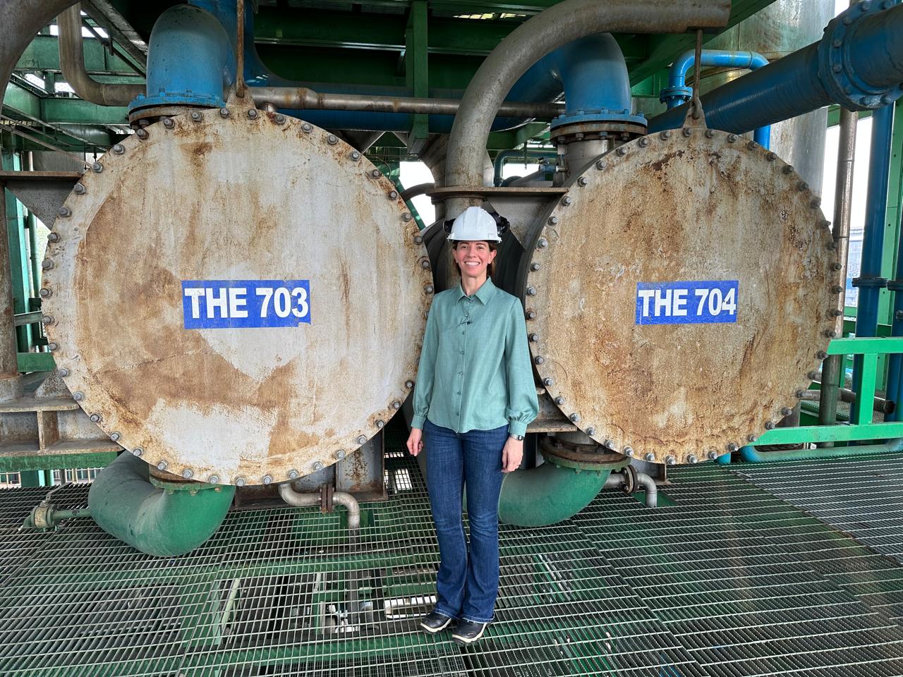 Gabriela da Silva em planta industrial, ao lado de trocadores de calor THE 703 e THE 704